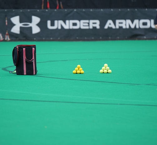 A sports field features a black and pink sports bag, two pyramids of yellow balls, and a portion of a person in dark athletic gear. The background displays a banner with the Under Armour logo.