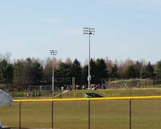 A sports field with a group of people engaging in athletic activities in the background. There is sports equipment like a soccer goal and a scoreboard visible. A chain-link fence runs across the foreground, and a statue or sculpture resembling an eagle is also seen. Trees and tall floodlights are positioned behind the field.