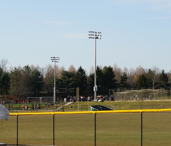 A sports field with a group of people engaging in athletic activities in the background. There is sports equipment like a soccer goal and a scoreboard visible. A chain-link fence runs across the foreground, and a statue or sculpture resembling an eagle is also seen. Trees and tall floodlights are positioned behind the field.