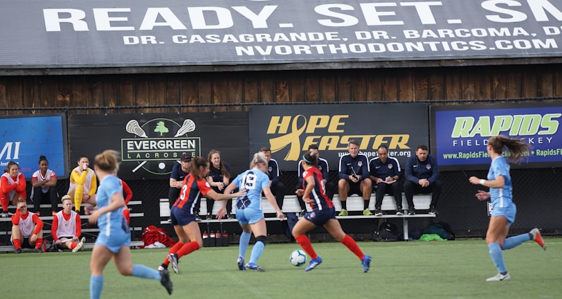Players engage in a competitive soccer game on a field while others are seated on the bench watching attentively. The action is taking place in front of a wooden structure displaying multiple advertisements, including orthodontics, lacrosse, and field hockey.