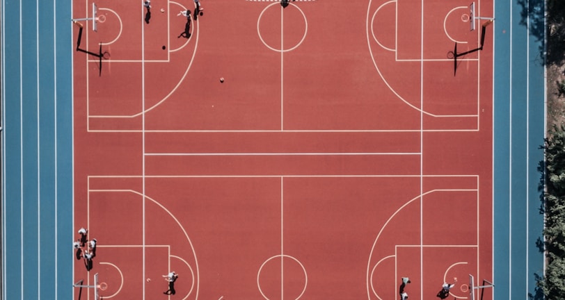 An aerial view of a large, vibrant sports complex featuring a red basketball court with multiple hoops and a surrounding blue running track. Several people are scattered across the area, engaged in various activities.
