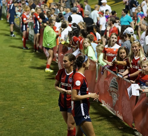 The image features a lively gathering of young soccer players and fans next to a field. Several female athletes in red and blue uniforms interact with an enthusiastic crowd standing behind a barrier. Some people in the crowd are holding objects, like pens and paper, possibly for autographs.