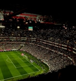 A large, illuminated stadium filled with a vast audience. The field is marked for soccer, with players actively participating in a match. The crowd is engaged, with numerous lights visible in the stands, possibly from phones. The stadium's roof structure is visible with red beams, and an electronic scoreboard shows a score of 0-0.