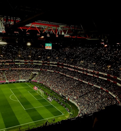 A large, illuminated stadium filled with a vast audience. The field is marked for soccer, with players actively participating in a match. The crowd is engaged, with numerous lights visible in the stands, possibly from phones. The stadium's roof structure is visible with red beams, and an electronic scoreboard shows a score of 0-0.