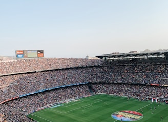 An expansive soccer stadium filled with thousands of spectators. The green field in the center features players and markings, surrounded by vibrant stands and advertising boards on the upper sides of the stadium. The audience is actively watching the game.