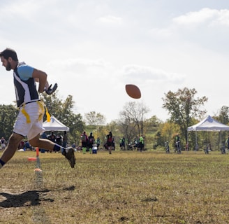 A person is engaged in a sports activity, running on a grassy field. The person is wearing a sports jersey and shorts. They are positioned in mid-air as they run, and a football is visible in front of them. In the background, there are several people along with tents and trees, indicating an outdoor sports event.