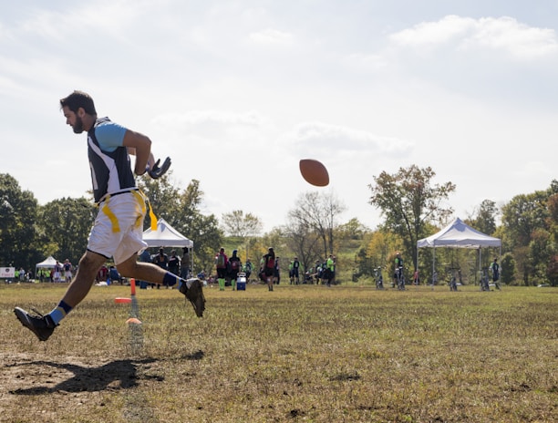 A person is engaged in a sports activity, running on a grassy field. The person is wearing a sports jersey and shorts. They are positioned in mid-air as they run, and a football is visible in front of them. In the background, there are several people along with tents and trees, indicating an outdoor sports event.