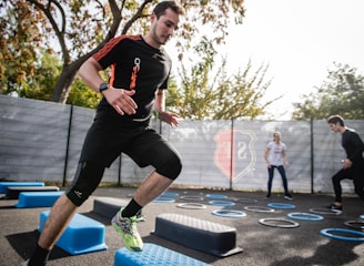 A man is actively participating in an outdoor fitness session, jumping over blue platforms with agility. Two other people in the background are also engaging in exercise, with rings scattered on the ground. The area is fenced and surrounded by trees, suggesting a park or recreational setting.