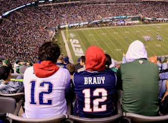 A crowded football stadium with fans sitting in the stands, wearing jerseys and hoodies. The field is visible in the background with teams lined up for a play. The audience is engaged, capturing the excitement of the game.