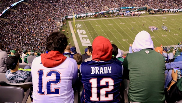 A crowded football stadium with fans sitting in the stands, wearing jerseys and hoodies. The field is visible in the background with teams lined up for a play. The audience is engaged, capturing the excitement of the game.
